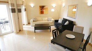 Una sala de estar con un sofá, una mesa de centro y una mesa de comedor con sillas. Hay un cuadro en la pared y un espejo sobre el sofá.