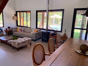 A living room with a sofa, ottoman, and dining table with chairs. There are large windows with views of greenery outside.