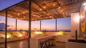 Un espace extérieur aménagé en terrasse, doté d’un toit de chaume, de guirlandes lumineuses et de tabourets de bar.