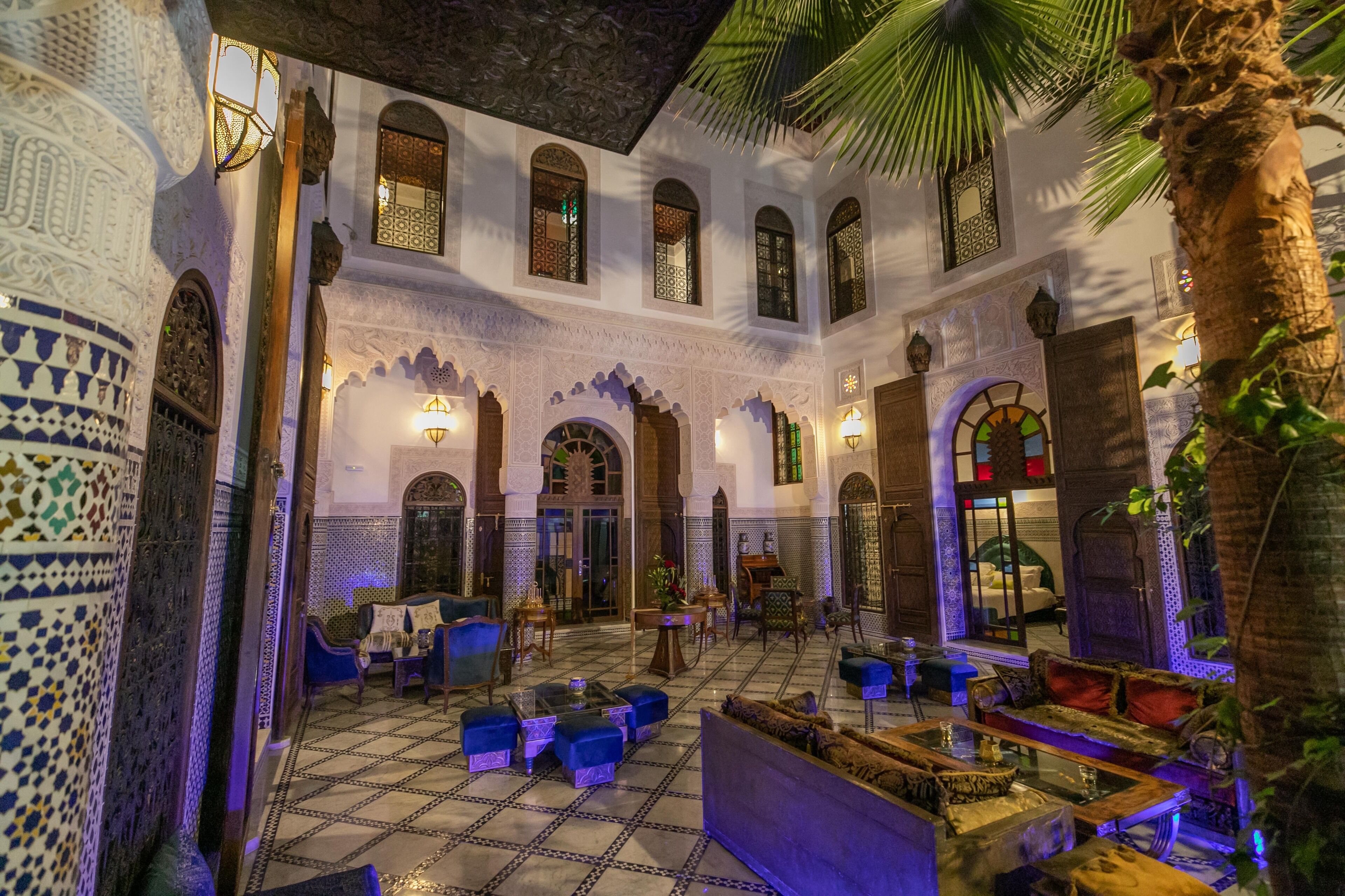 A traditional Moroccan interior with ornate tile work, arched doorways, and a central seating area with sofas and tables.