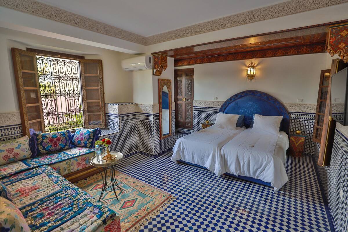 A bedroom with a checkered floor, a large bed with white bedding, a sofa with colorful cushions, and a small table with a vase.