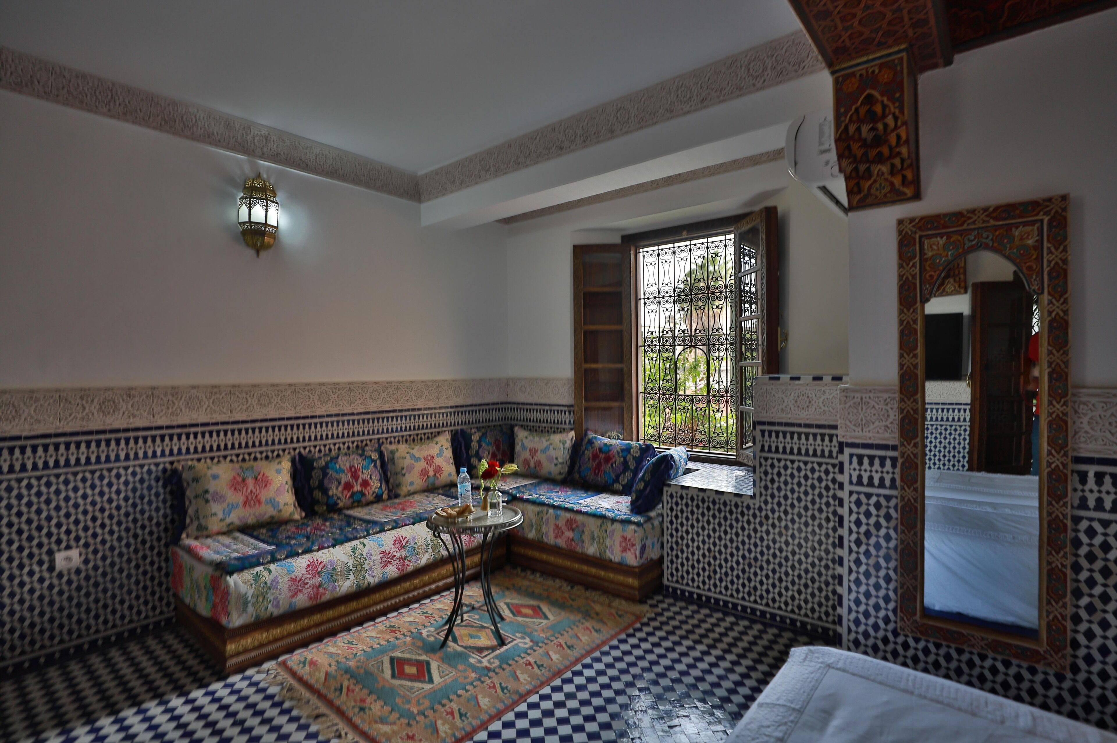 A room with a patterned sofa, a small round table with a vase, a large mirror, and a window with metal bars.