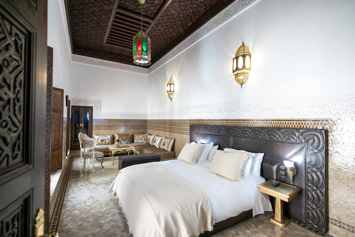 A spacious bedroom with a large bed, a sitting area with a sofa and chairs, and decorative wall sconces.