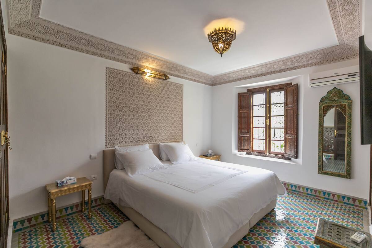 A bedroom with a large bed, a nightstand, a wall-mounted lamp, a window with wooden shutters, and a decorative mirror.