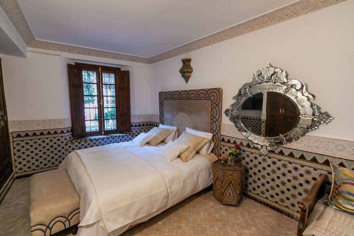 A bedroom with a large bed, a decorative mirror, and a window with wooden shutters.
