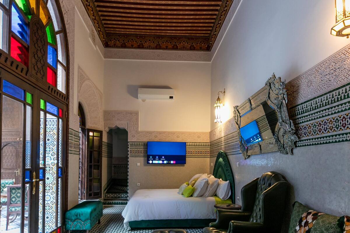 A room with a bed, a green sofa, a TV, and a tray with a bottle and a flower.
