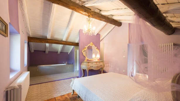 Une chambre avec un plafond en bois, un lustre, un lit recouvert d’une courtepointe blanche, une coiffeuse avec un miroir et une vue sur la salle de bain.