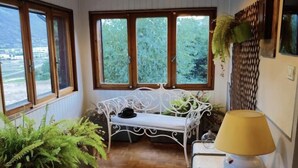 Une pièce chaleureuse avec un plancher en bois, un banc blanc et de grandes fenêtres donnant sur un paysage verdoyant.
