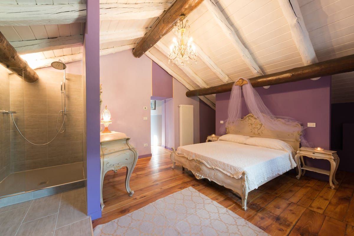 A bedroom with a wooden ceiling, a large bed with a canopy, a bedside table with a lamp, and a shower area.