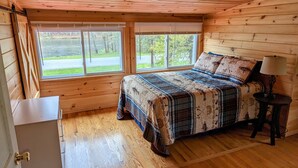 A cozy cabin bedroom with a wooden bed, a small bedside table, and a window overlooking a road and greenery. - Waterfront house on the upper most portion of the West Branch Delaware. (Deposit)