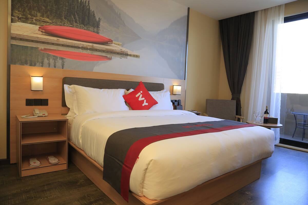 A hotel room with a large bed, a nightstand, a small table with a wine bottle, and a view of a mountainous landscape through the window.