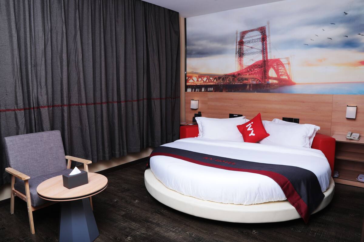 A hotel room with a round bed, a chair, a small table, a painting of a bridge, and a curtain.