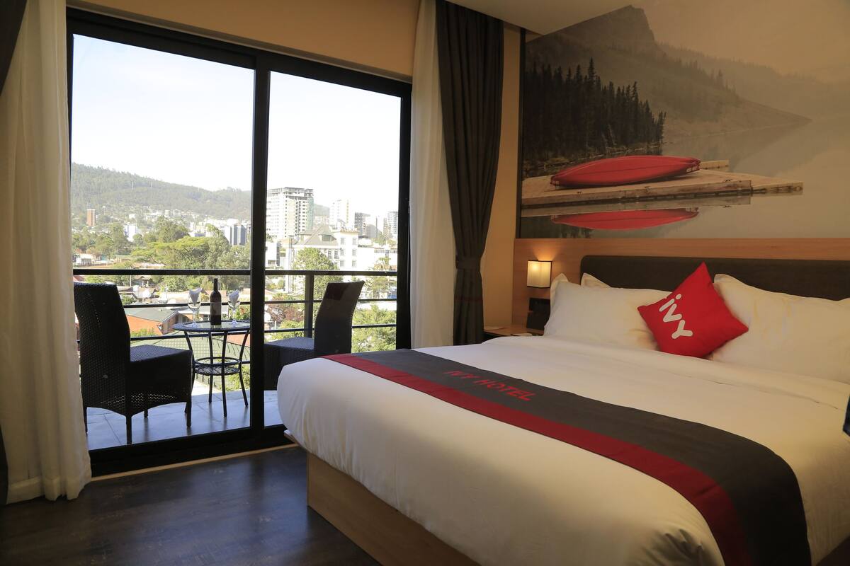 A hotel room with a large bed, a balcony with a table and chairs, and a city view.