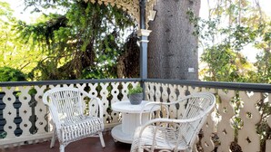 Un espace extérieur aménagé avec des sièges, protégé par un toit, comprenant des chaises en osier blanc et une table ronde, le tout entouré d’une clôture décorative et d’un grand arbre.