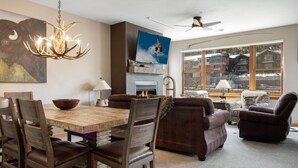 A dining area with a wooden table and chairs, a chandelier, a fireplace, and a television mounted on the wall.