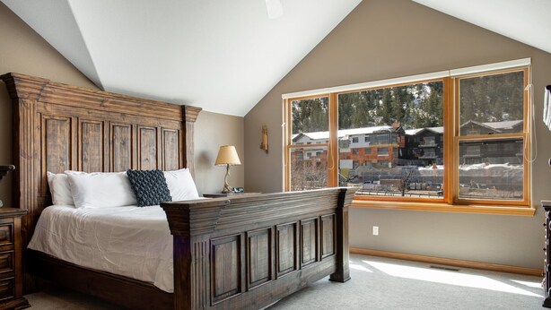 A bedroom with a wooden bed, a nightstand, a lamp, and a large window with a view of buildings and trees.