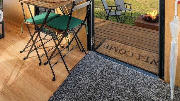 A modern interior with a wooden table and chairs, a view through large glass doors to an outdoor area, and a doormat with 'WELCOME' on it.