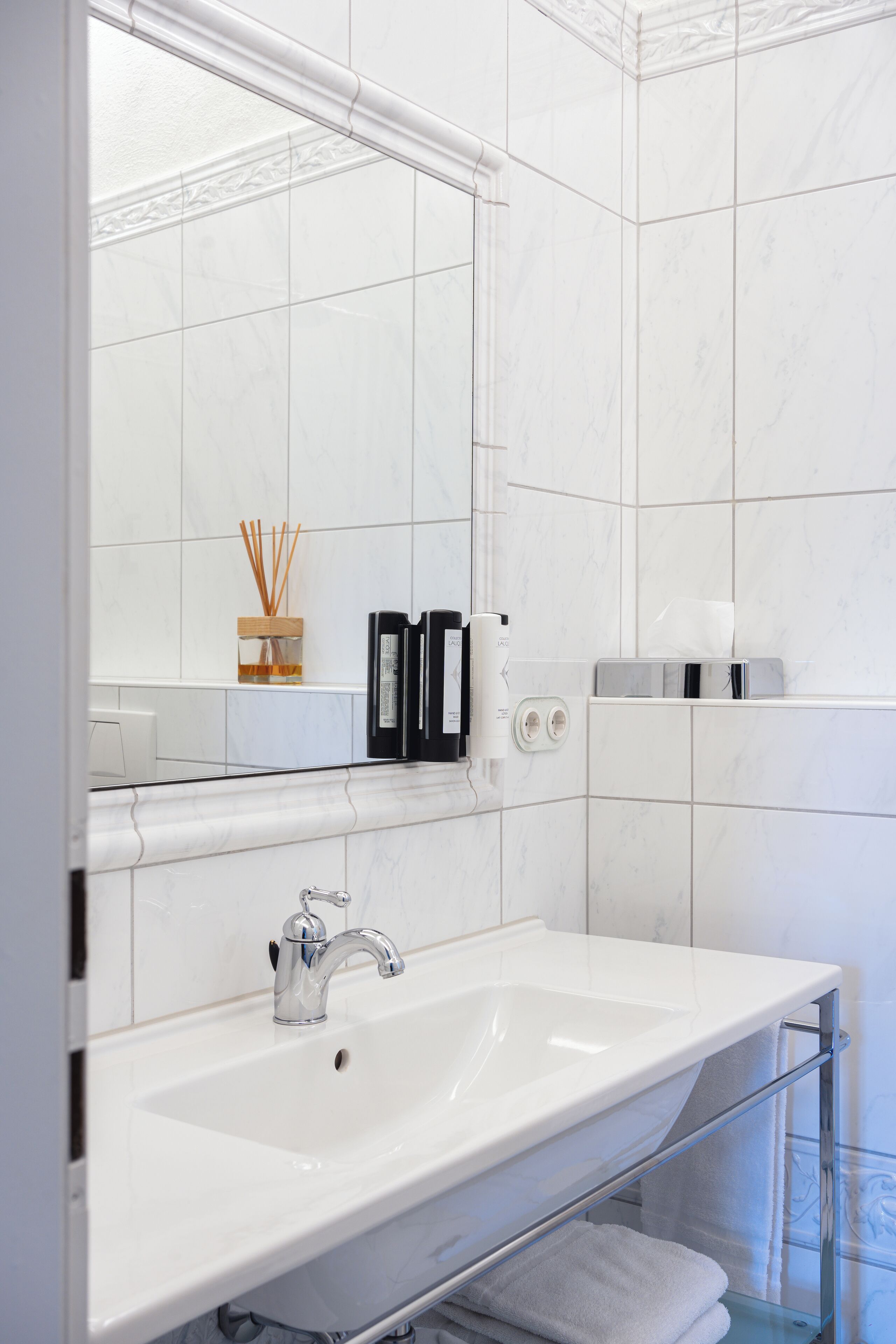 Un baño moderno con un lavamanos blanco, grifo cromado y un espejo encima. Hay dos botellas de jabón para manos y una caja de pañuelos en la pared.