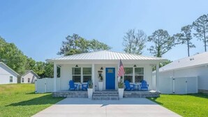Ein eingeschossiges Gebäude mit weißer Fassade und blauer Tür, inklusive einer kleinen Veranda mit blauen Stühlen und einer amerikanischen Flagge.