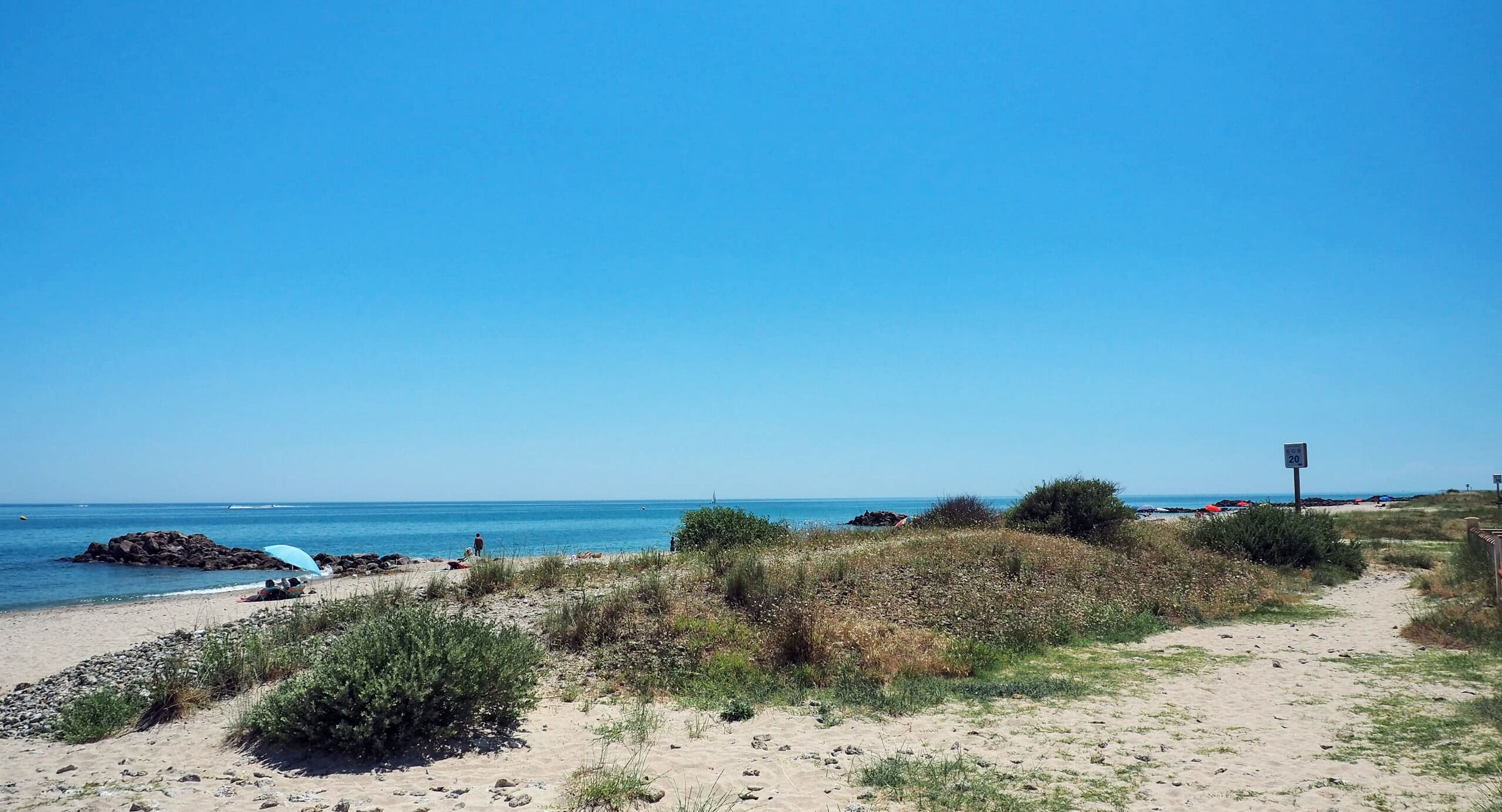 Una playa con cielo despejado, camino de arena y algo de vegetación.