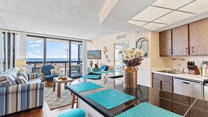 Ein modernes Wohnzimmer mit großem Fenster, einer Couch, einem Esstisch und einer Küchenzeile mit Holdekor und Spüle.