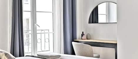 A hotel room with a bed, a desk, a chair, a large window with curtains, and a balcony.
