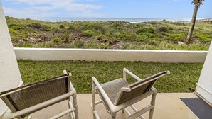 Ein Balkon mit zwei Korbstühlen, der auf einen Strand mit einer Palme und einem weißen Zaun blickt.