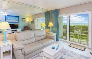 A modern living room with a beige sofa, a flat-screen TV, and a balcony with a view of the ocean. - Oceanfront Getaway (St. Augustine)