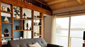 Une pièce chaleureuse avec un canapé d’angle, des étagères en bois et une grande fenêtre donnant sur une étendue d’eau.