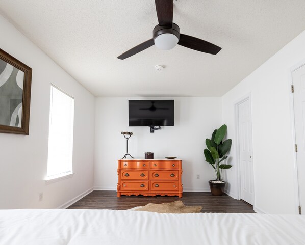 A bedroom with a bed, a ceiling fan, a dresser, a television, a window, and a plant.