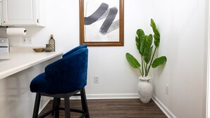 A kitchen with a white countertop, a blue chair, a framed abstract artwork, and a potted plant.