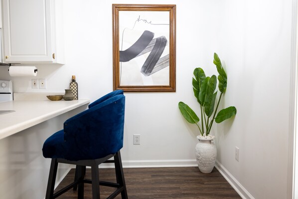 A kitchen with a white countertop, a blue chair, a framed abstract artwork, and a potted plant.