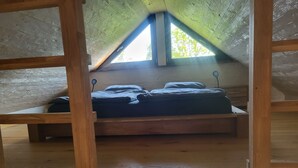 Une chambre à coucher aménagée dans un grenier, avec un plafond en pente, des poutres en bois, un lit avec des oreillers et une vue sur les arbres à travers une lucarne.
