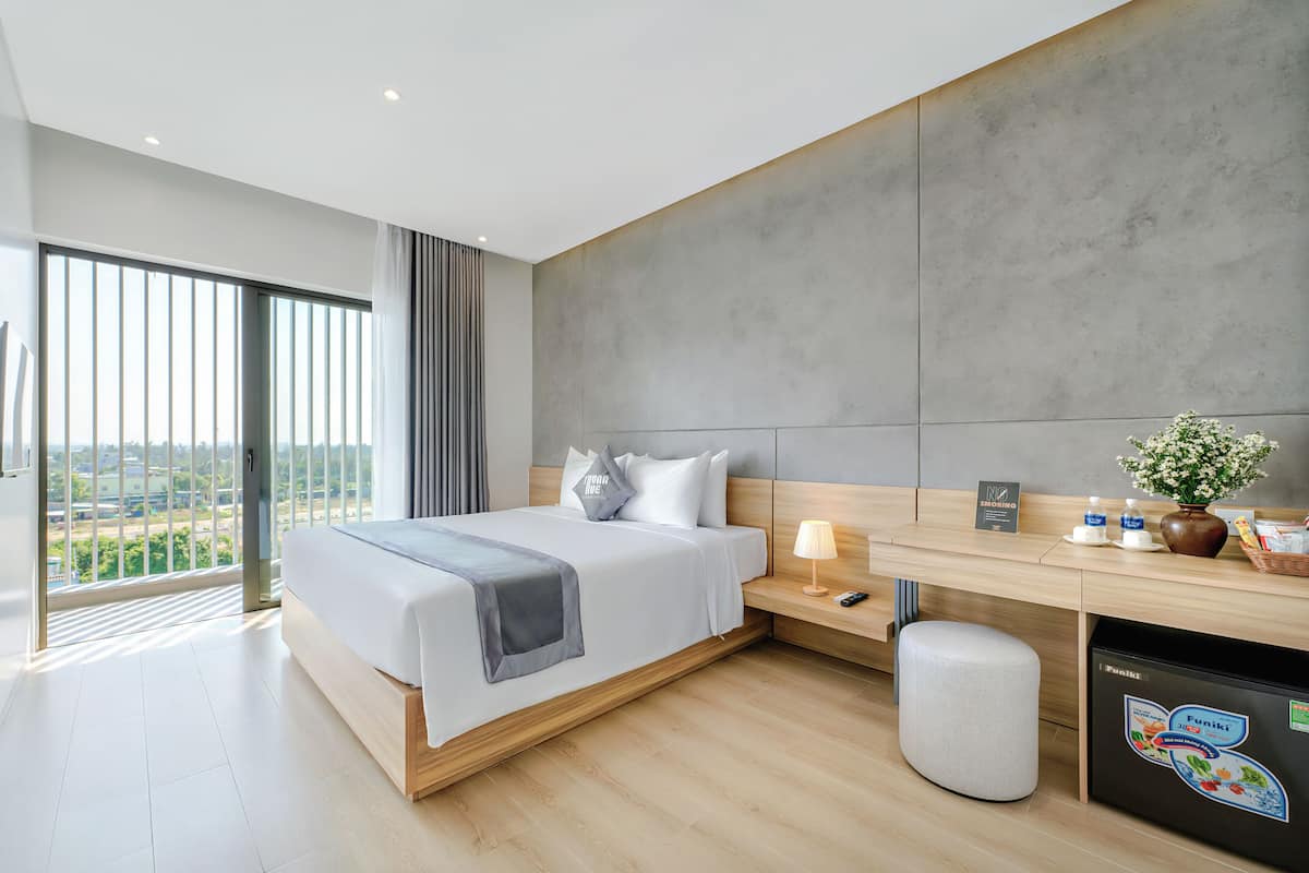 Quarto de hotel moderno com uma cama grande, uma escrivaninha, um banquinho pequeno, uma mesa de cabeceira e vista para a cidade.