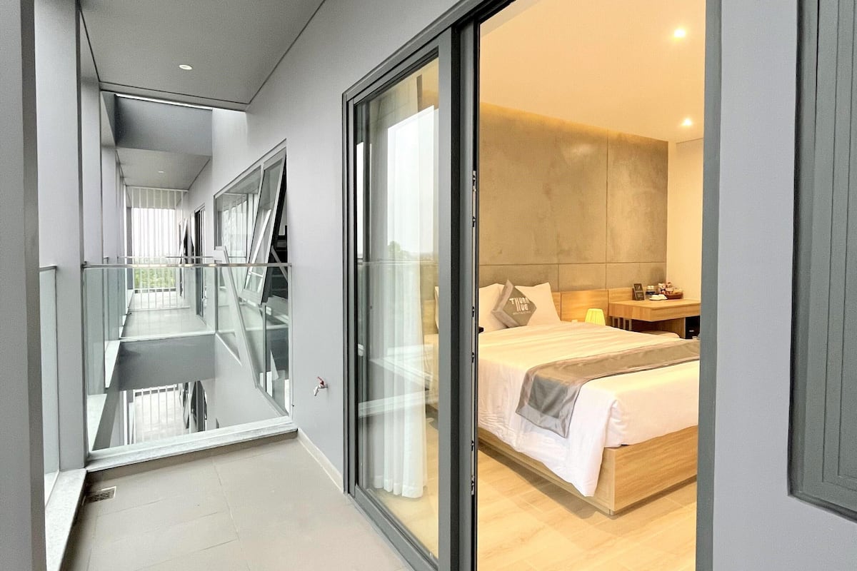 Corredor moderno de hotel com porta de vidro que dá para um quarto com cama, mesa de cabeceira e vista para a cidade.