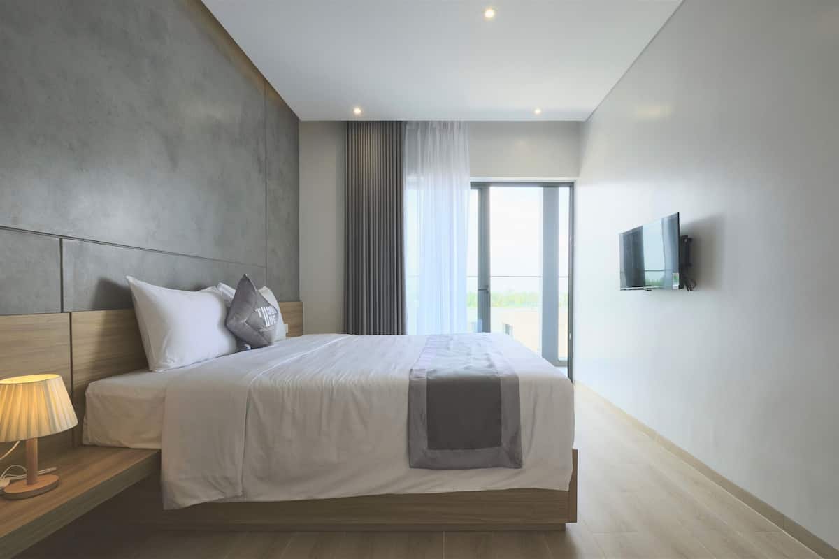 Quarto de hotel moderno com uma cama grande, uma mesa de cabeceira com abajur, televisão na parede e vista para o exterior pela janela.