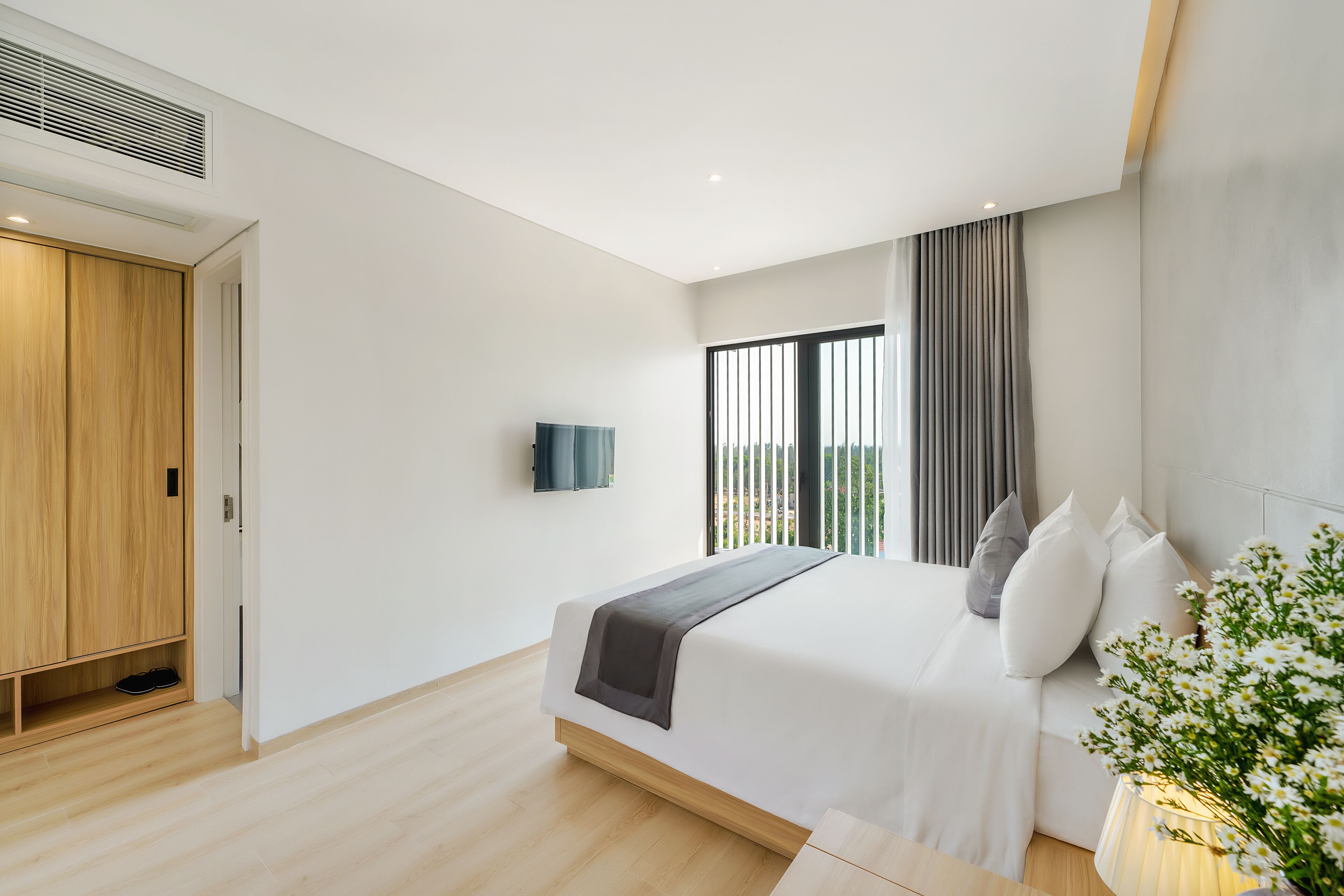 A modern bedroom with a large bed, a TV mounted on the wall, and a view of the outdoors through a window.