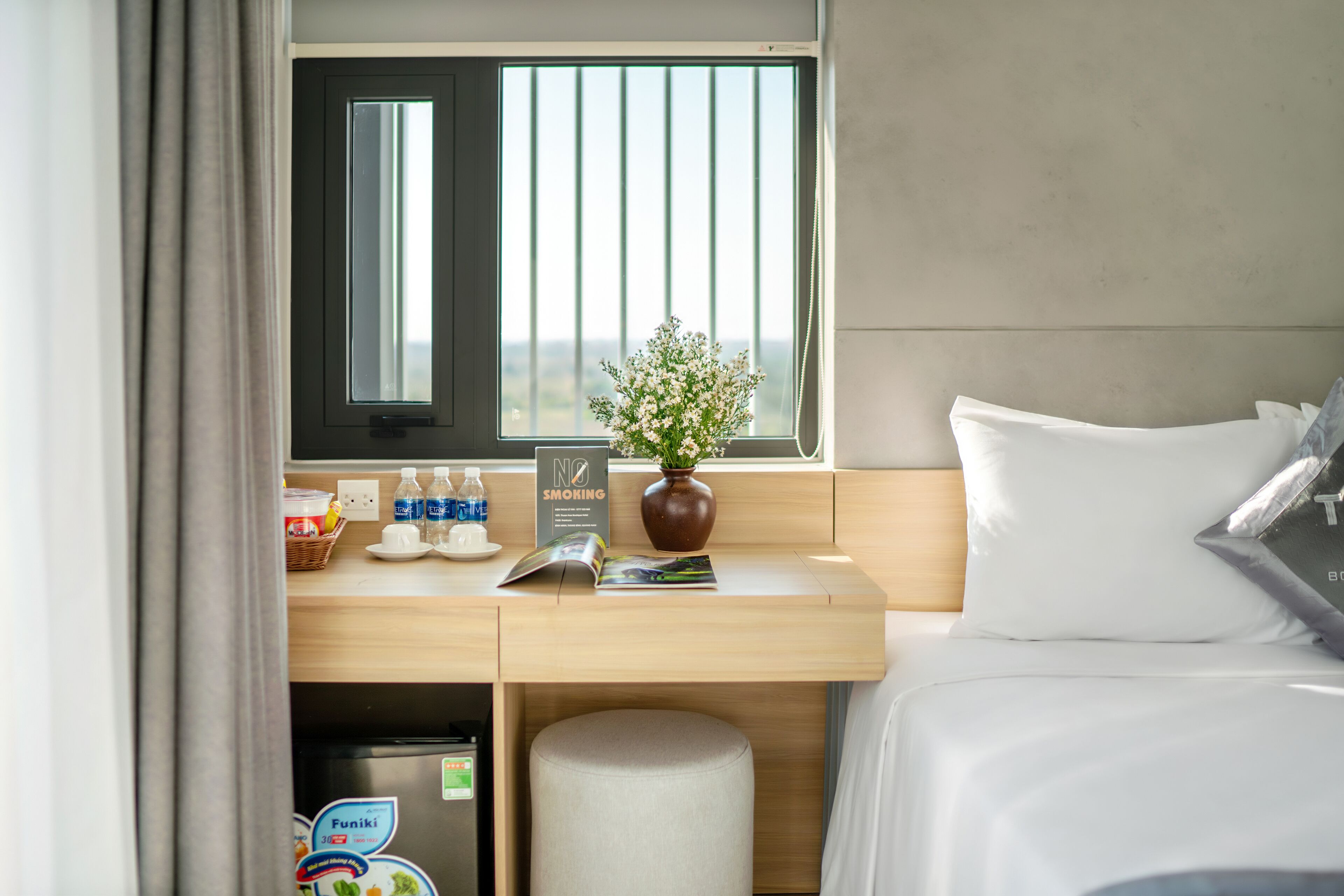 A hotel room with a bed, a desk with a vase of flowers, a window with blinds, and a sign prohibiting smoking.