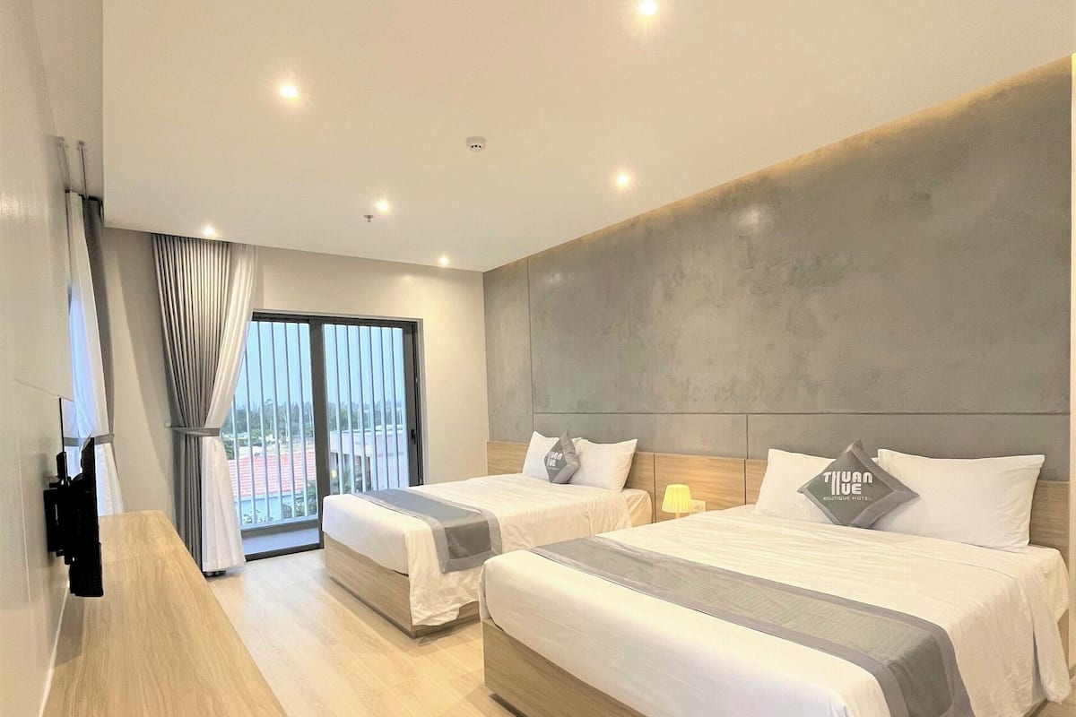 Quarto de hotel moderno com duas camas, uma secretária de madeira e uma janela grande com cortinas.