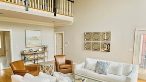 Un salon spacieux avec un canapé blanc, un fauteuil en cuir marron et une table basse en verre. Un escalier mène à un niveau supérieur, et une porte donne accès à une autre pièce.