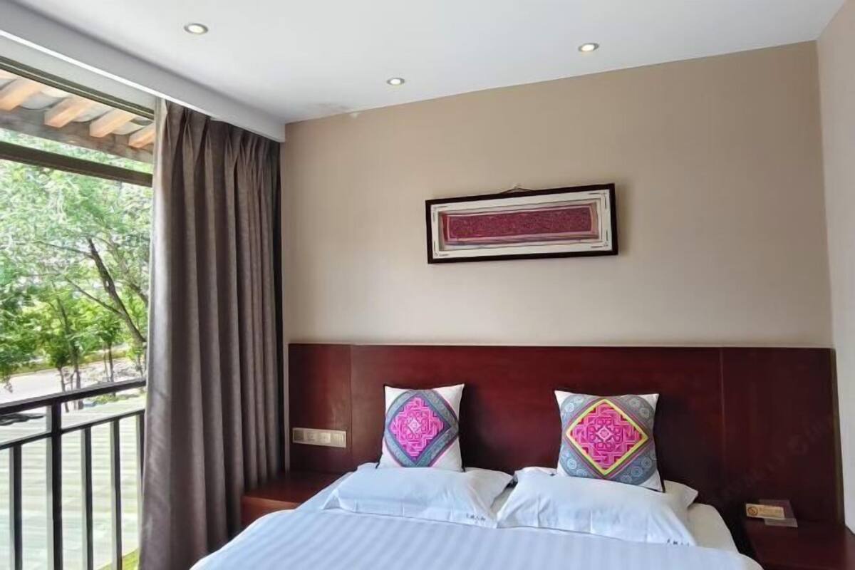 A hotel room with a bed, a window with curtains, a framed picture on the wall, and a view of trees outside.