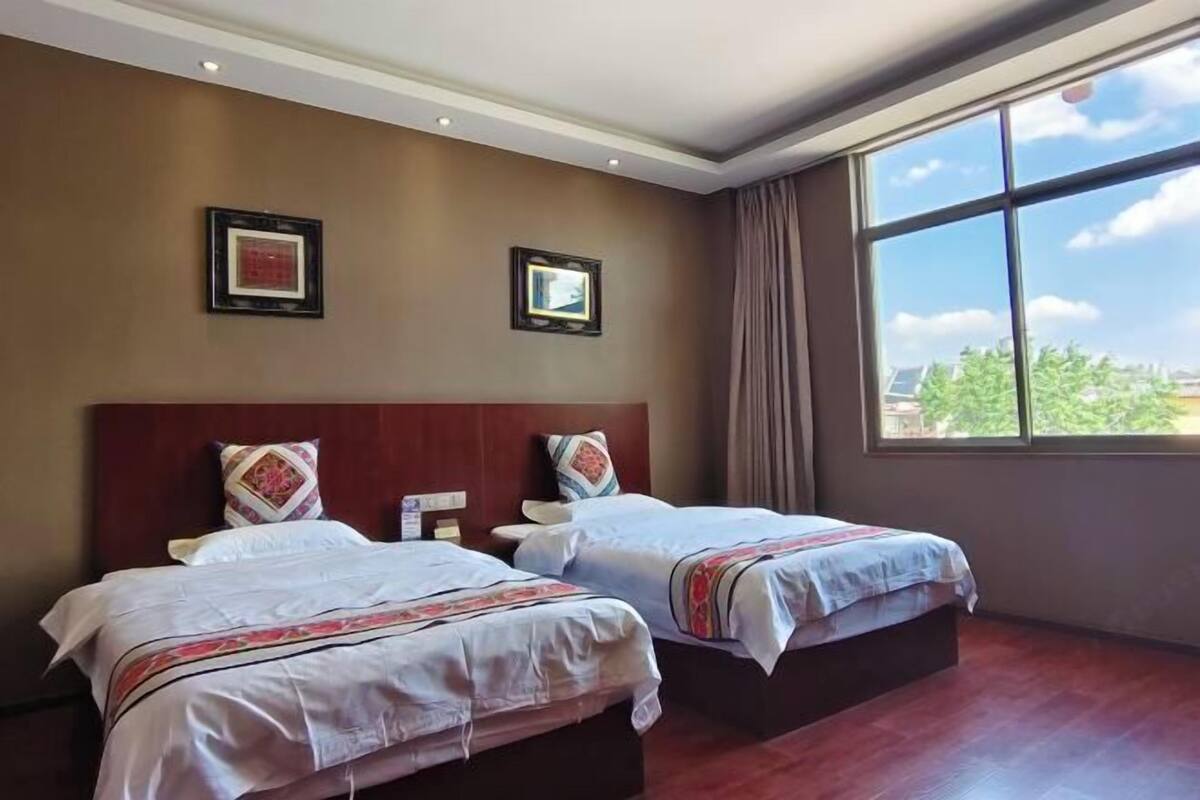 A hotel room with two beds, a large window, and a view of greenery outside.