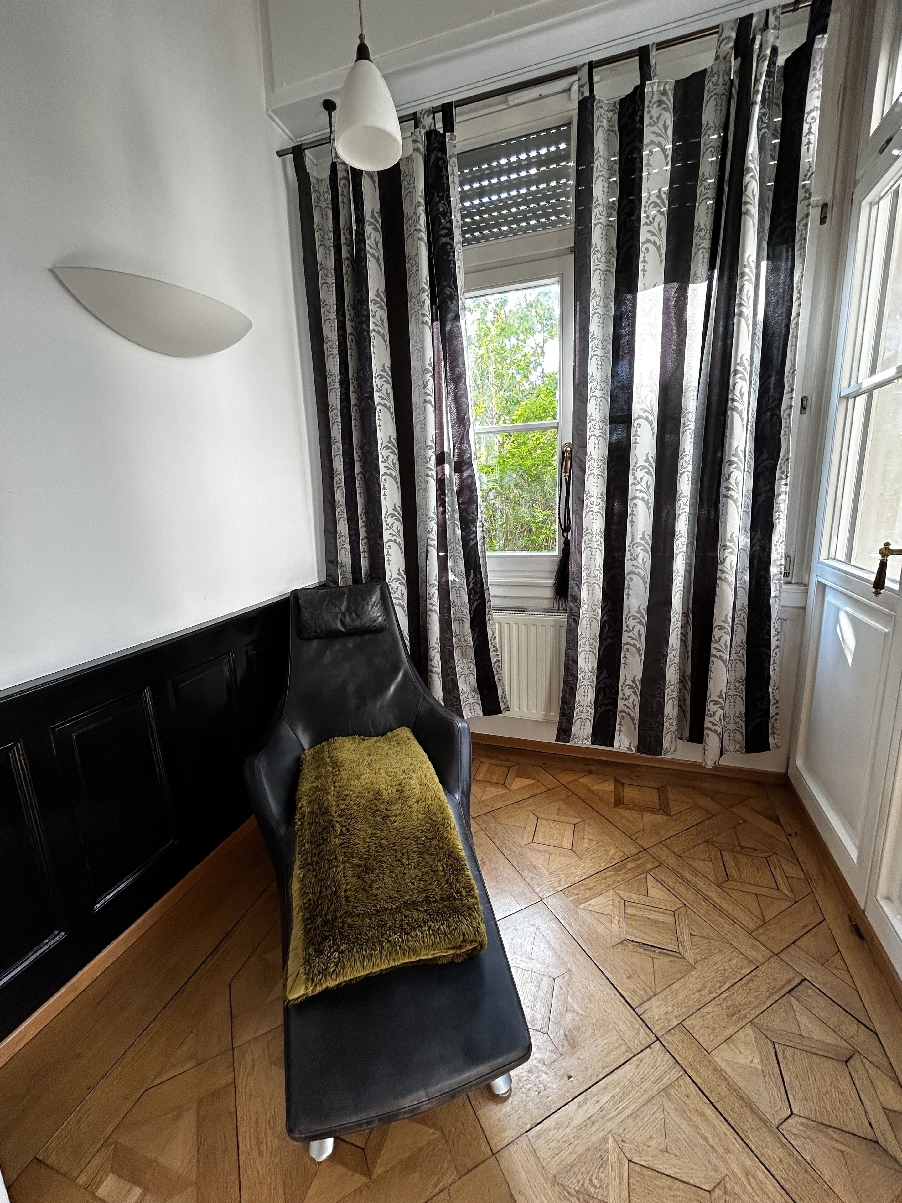 A room with a black leather chair, a small table with a green cushion, a hanging light fixture, and a window with patterned curtains.