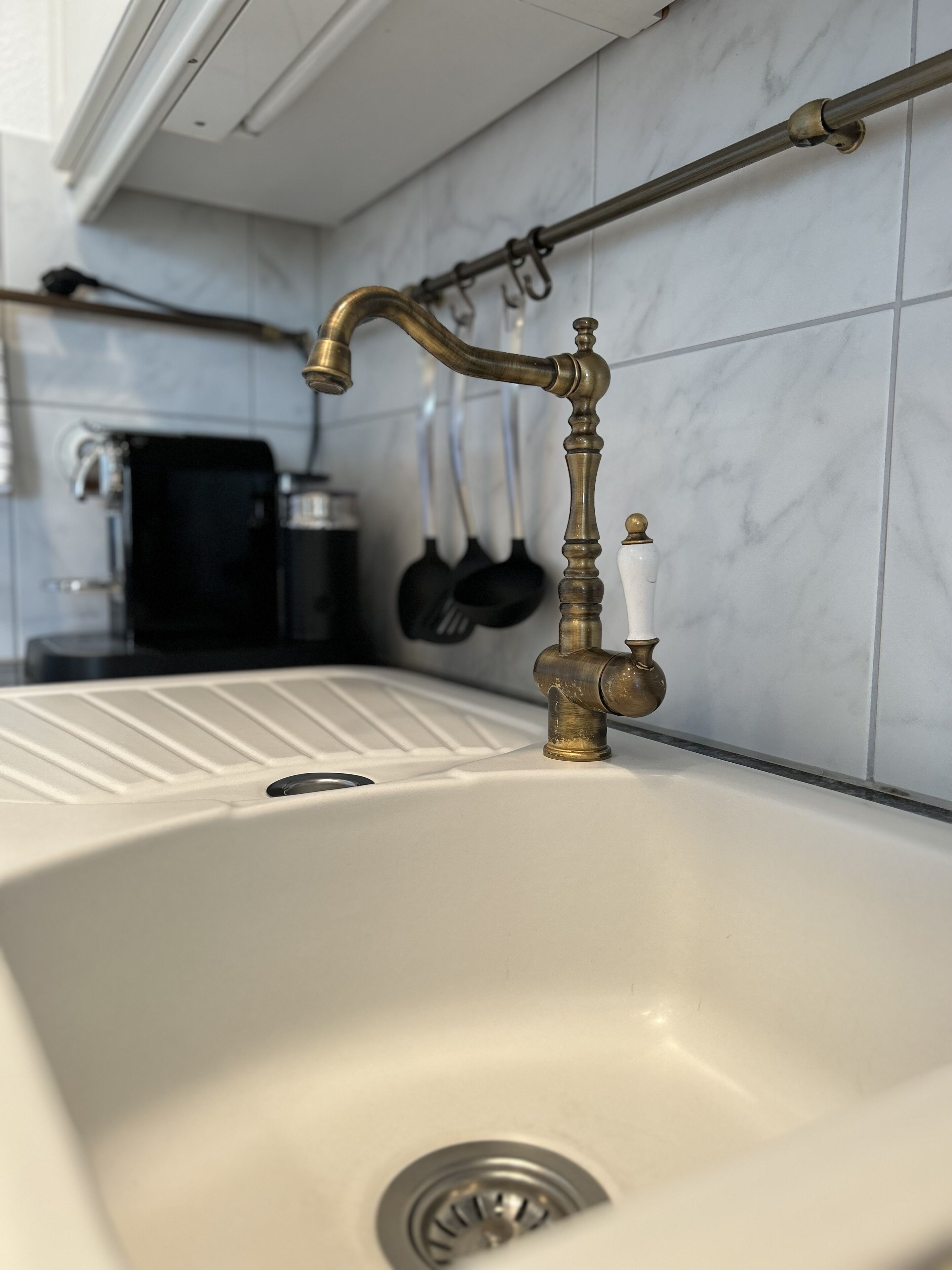 A kitchen sink with a vintage faucet, a black coffee maker, and a set of kitchen utensils on the wall.