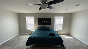 Une chambre à coucher comprenant un lit, un ventilateur de plafond, deux fenêtres équipées de stores et une œuvre d’art accrochée au mur.