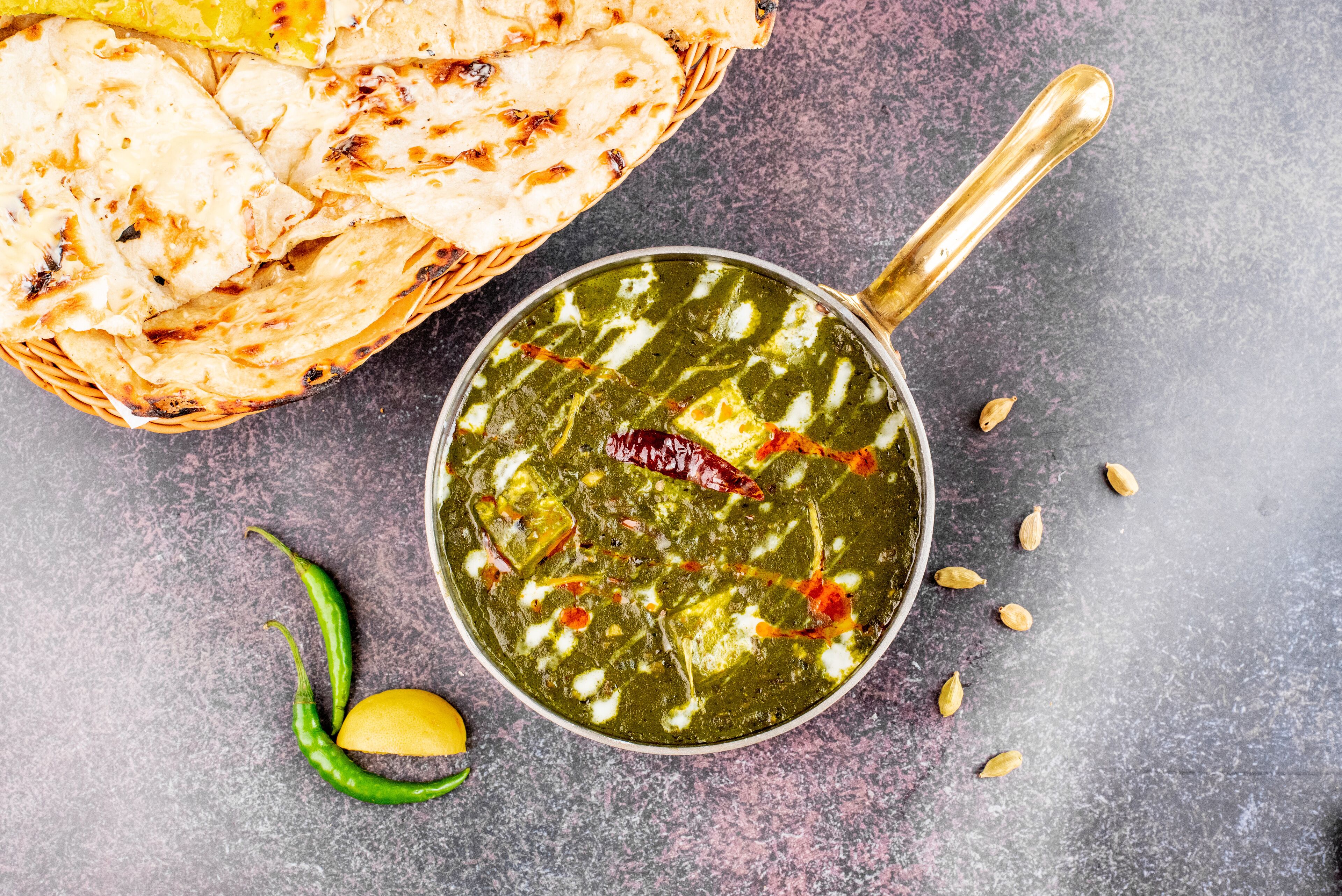 A bowl of green curry with paneer, accompanied by naan bread and a lemon.