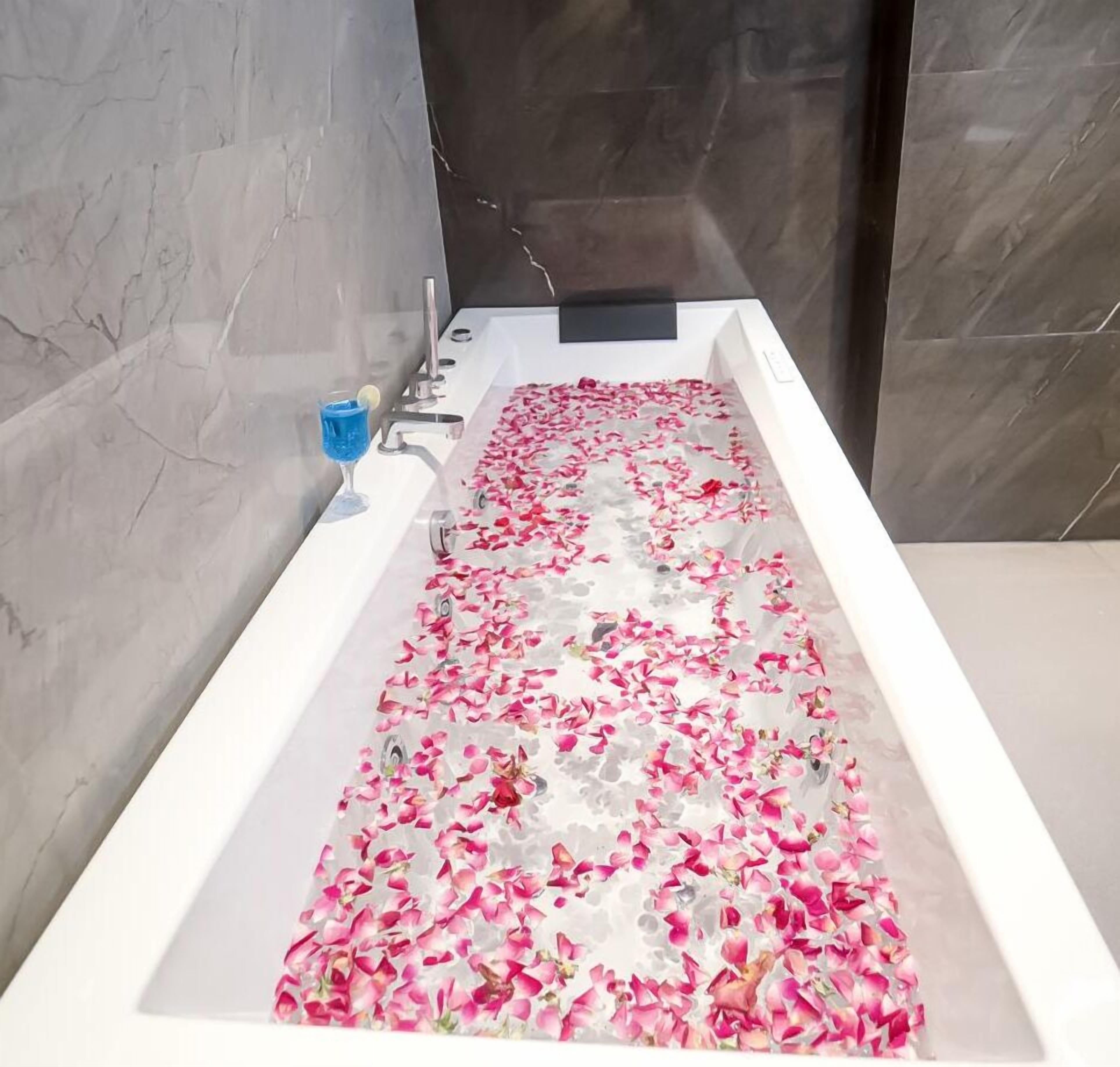 A bathtub filled with rose petals and a blue drink on the side.