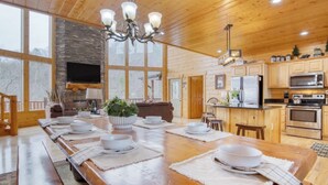 A spacious dining area with a long wooden table set for a meal, a stone fireplace, wooden cabinets, and a kitchen area with modern appliances. - Luxurious 4BD Lodge with HOT TUB! Fireplace! (Sevierville)