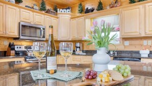 A kitchen with wooden cabinets, a microwave, a wine bottle, two glasses of wine, a vase with flowers, and a cheese board with grapes and crackers. - Luxurious 4BD Lodge with HOT TUB! Fireplace! (Sevierville)