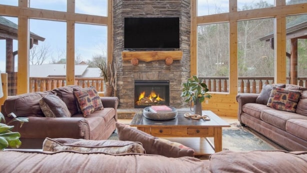 A cozy living room with a stone fireplace, leather sofas, and a large window overlooking a deck. - Luxurious 4BD Lodge with HOT TUB! Fireplace! (Sevierville)